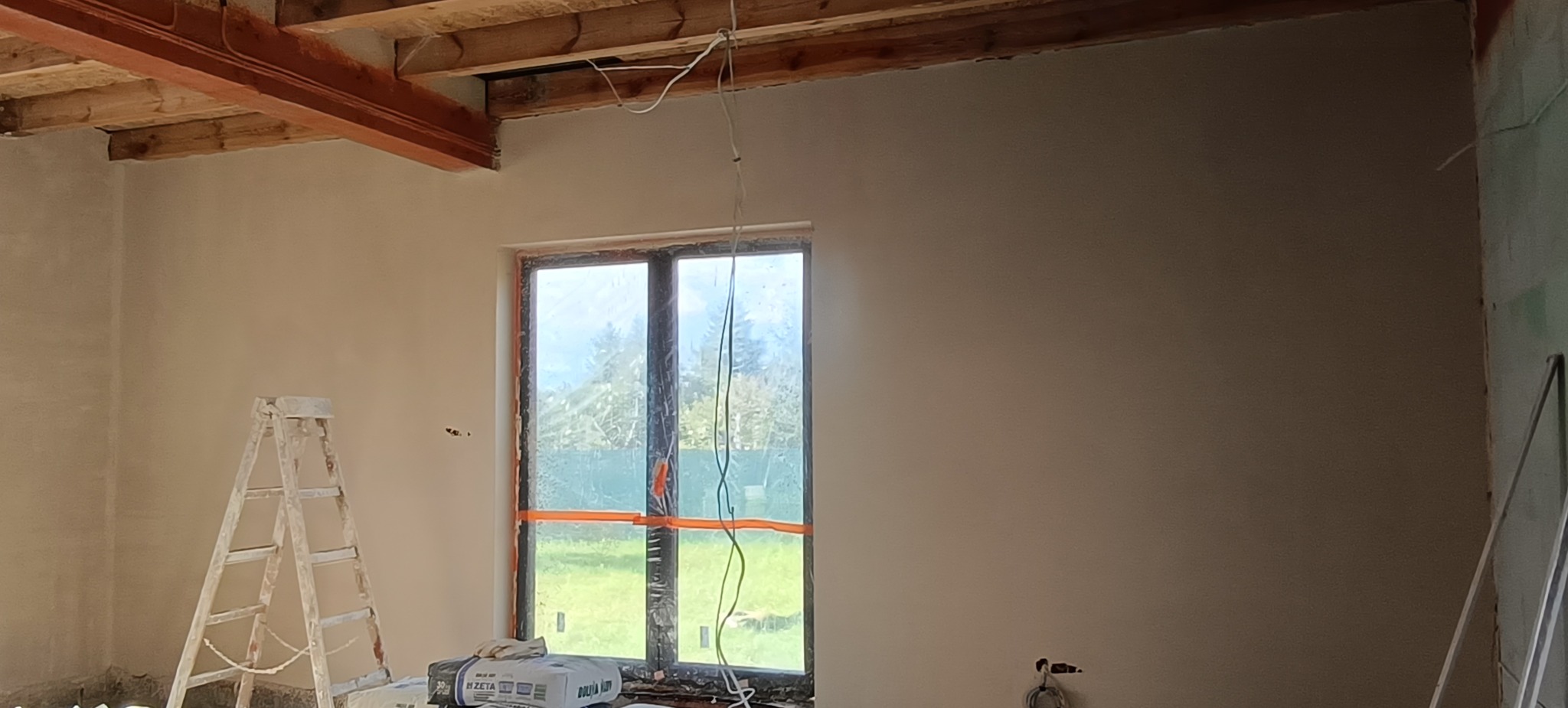 Wnętrze po nałożeniu gładzi, widoczne drewniane belki stropowe, okno zabezpieczone folią, drabina malarska i worki z zaprawą. Ściany gładkie, przygotowane do dalszych prac.