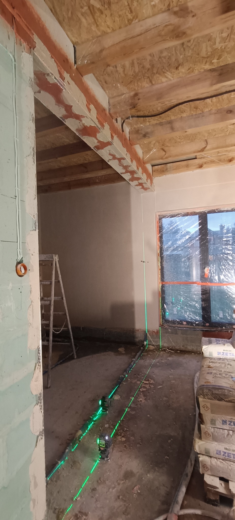 Wnętrze w trakcie prac wykończeniowych. Widoczne ściany po szpachlowaniu, laser krzyżowy wyznaczający poziom, worki z zaprawą i drabina. Surowe drewno na suficie.