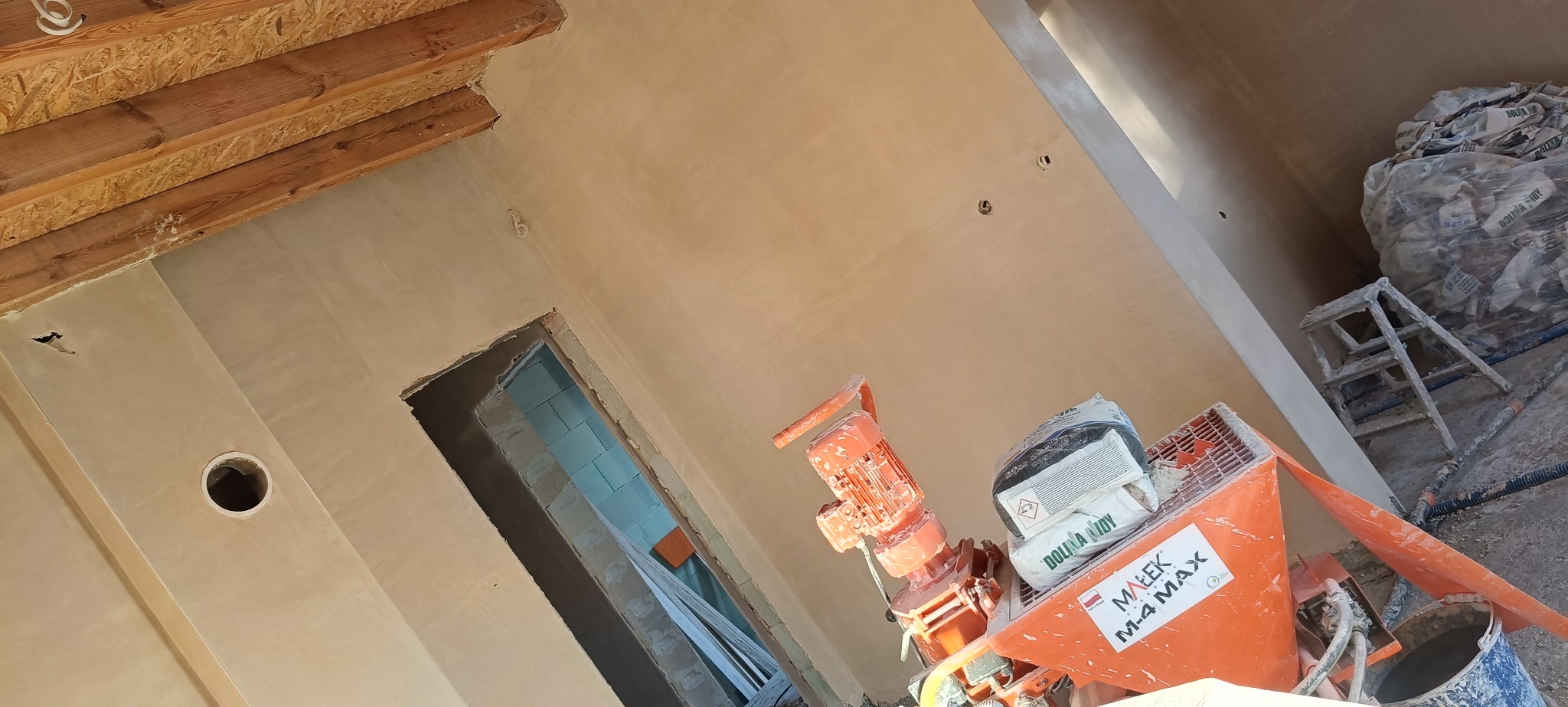 Świeżo otynkowane ściany wewnątrz budynku z widoczną maszyną tynkarską Malek M-4 MAX i workami z zaprawą. Widoczne drewniane elementy konstrukcyjne stropu.
