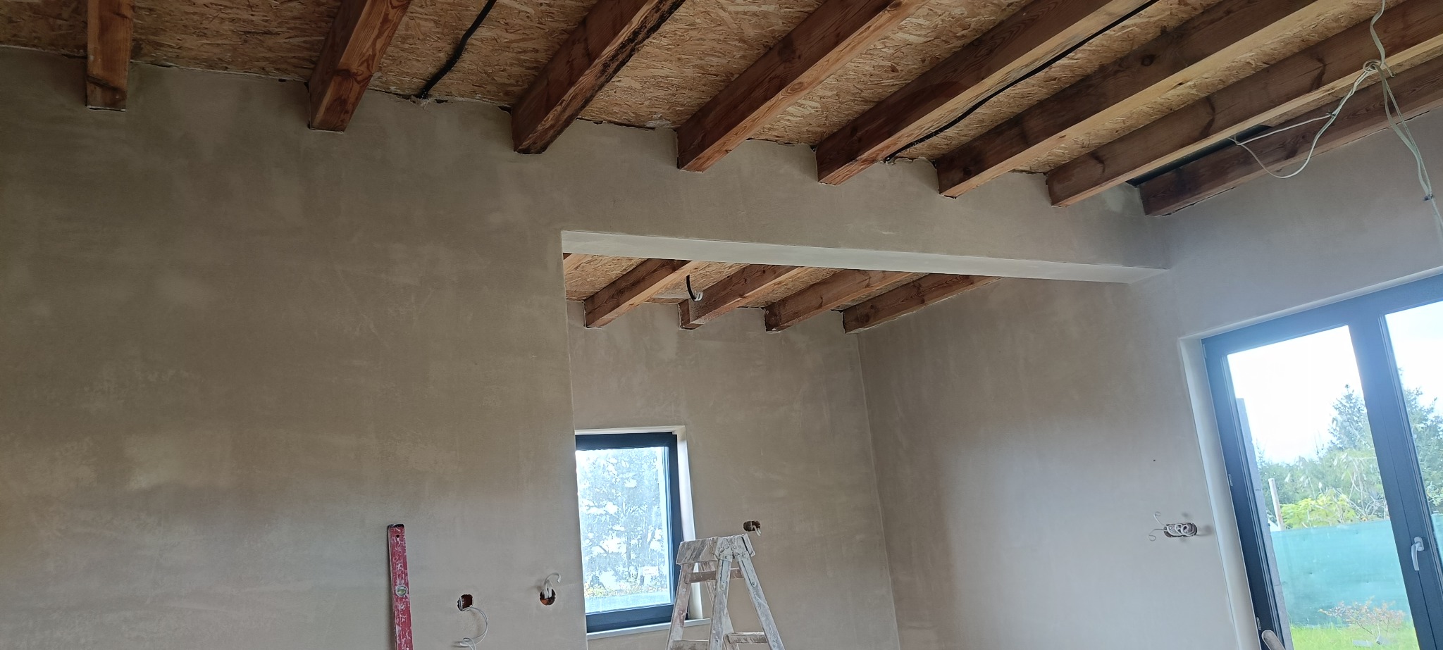 Surowe ściany po tynkowaniu maszynowym w jasnym wnętrzu z drewnianym stropem, widoczne okna i drabina malarska. Ściany przygotowane do dalszych prac wykończeniowych.