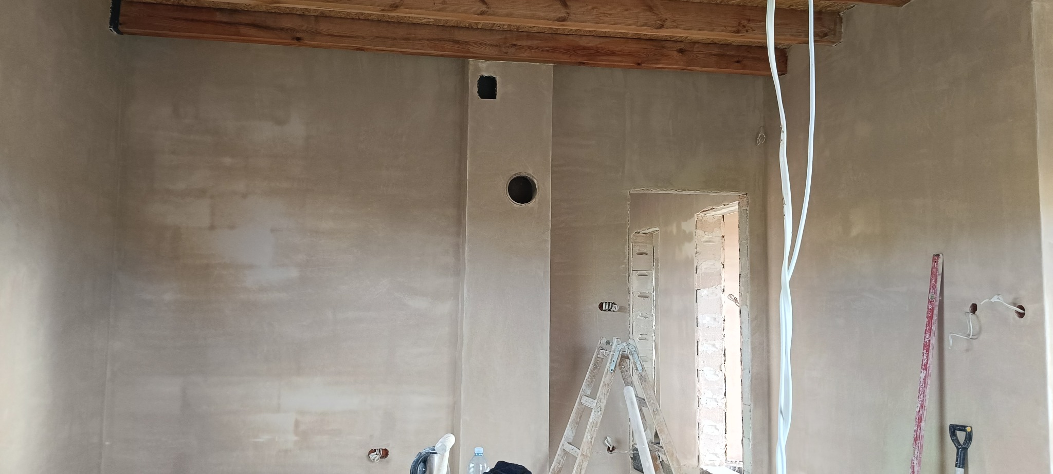 Wnętrze po tynkowaniu maszynowym: ściany pokryte świeżym tynkiem, widoczne otwory wentylacyjne, drabina i narzędzia. Surowy stan wykończenia, przygotowanie do dalszych prac.