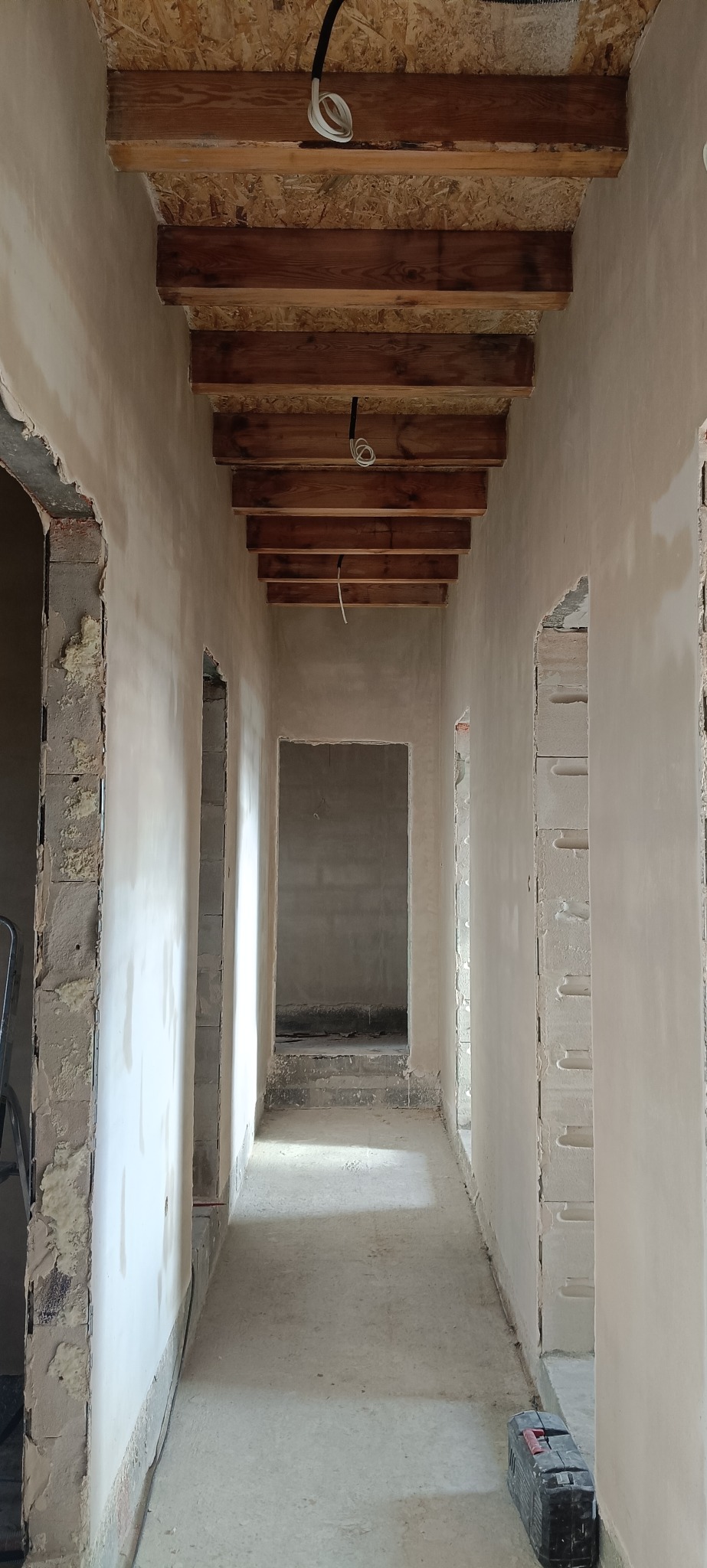 Korytarz w trakcie prac wykończeniowych: ściany pokryte świeżym tynkiem, widoczne drewniane elementy konstrukcyjne pod sufitem, surowy stan wykończenia.