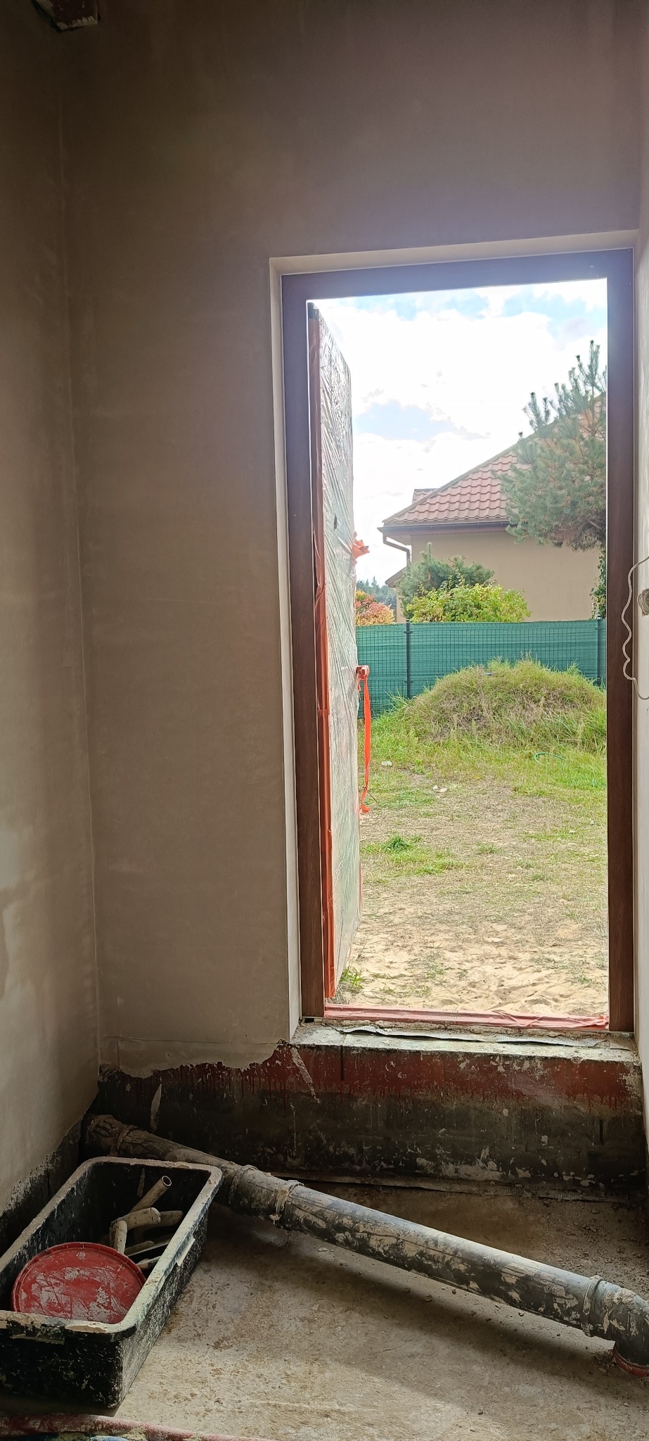 Wnętrze w trakcie prac wykończeniowych: ściany po tynkowaniu, zabezpieczone okno folią, widoczne narzędzia i resztki materiałów budowlanych na podłodze.