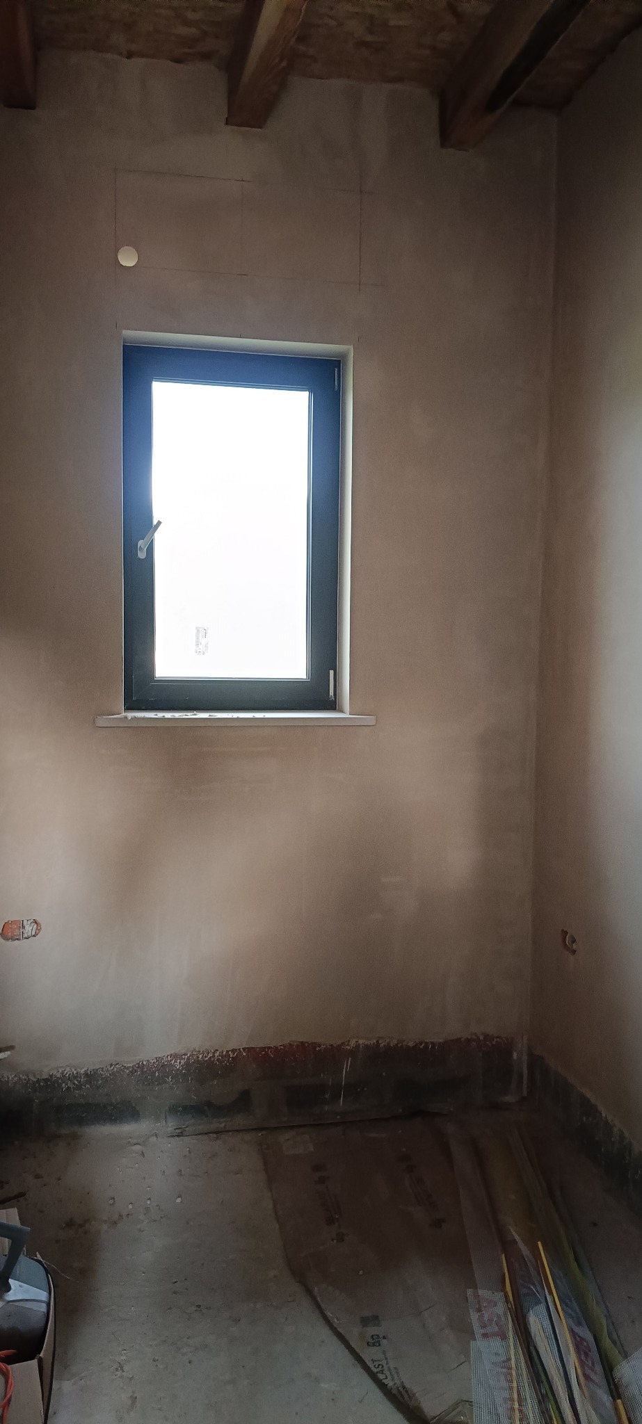 Pokój po nałożeniu tynku, widoczne ślady po instalacjach elektrycznych, okno z czarną ramą i drewniane belki stropowe. Ściany w kolorze beżowym.