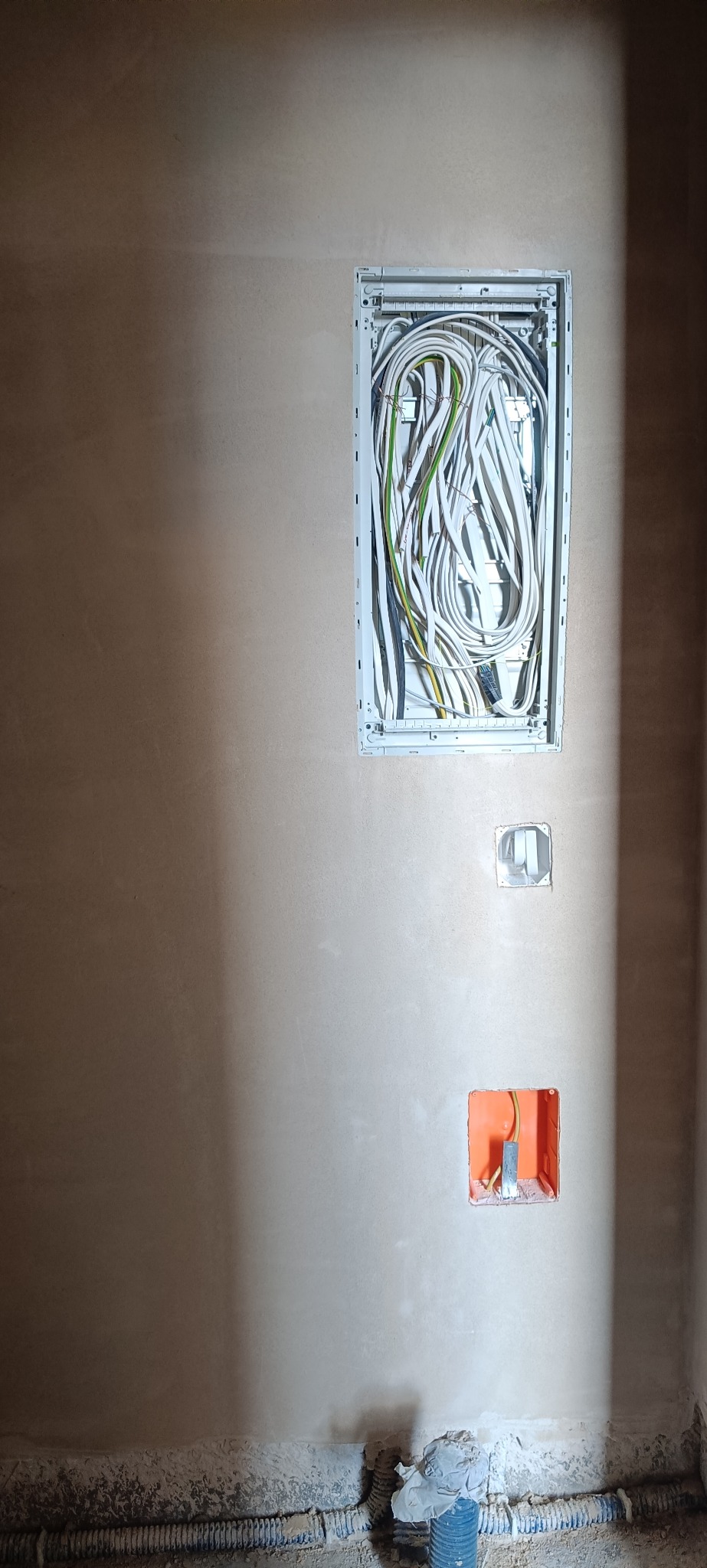Ściana po tynkowaniu z zamontowaną skrzynką elektryczną i puszkami pod gniazdka. Widoczne przewody elektryczne. Prace wykończeniowe w trakcie realizacji.