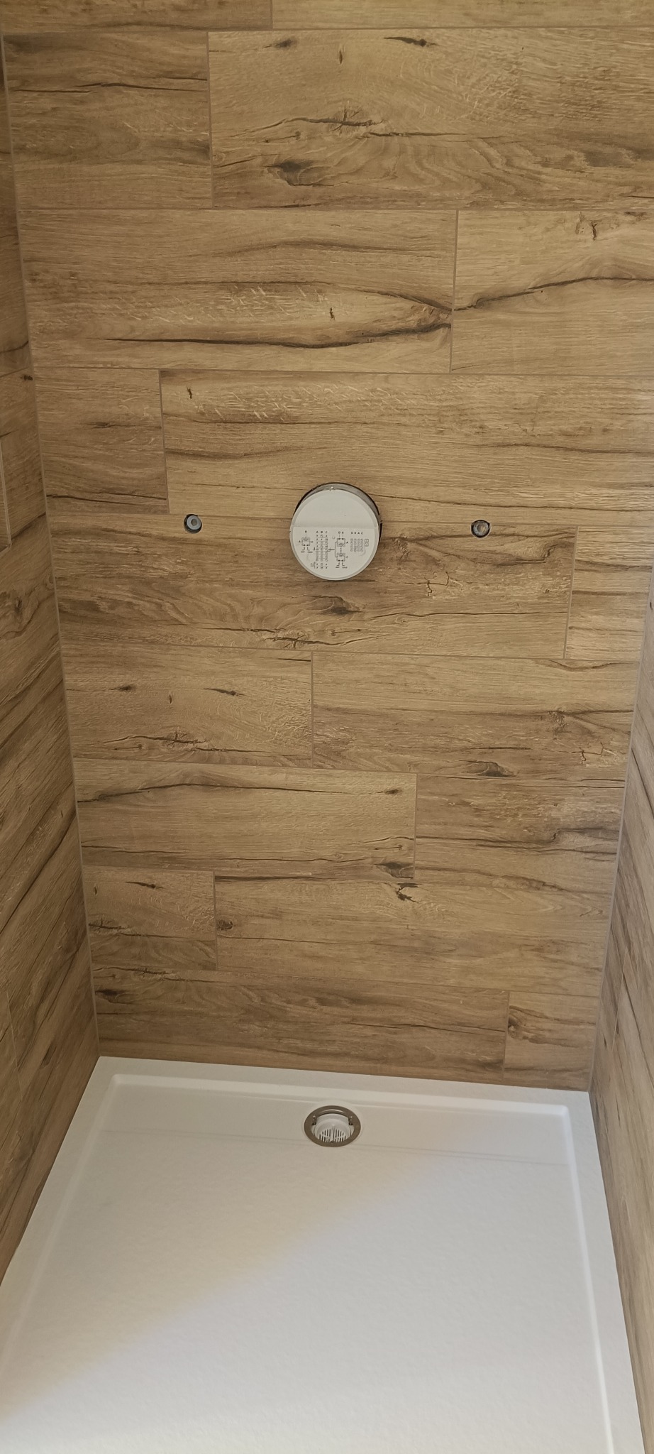 Wykończona kabina prysznicowa z drewnopodobnymi płytkami na ścianach i białą brodzikiem. Widoczny punkt świetlny na suficie i odpływ liniowy w brodziku.