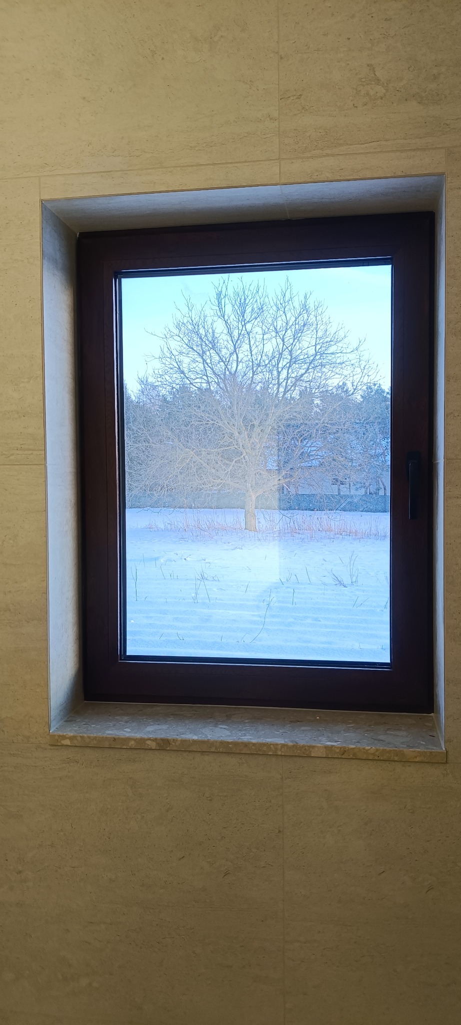 Okna z brązową ramą w jasnej, wykończonej płytkami wnęce. Widok na zimowy krajobraz z drzewem bez liści i śniegiem. Minimalistyczny design.