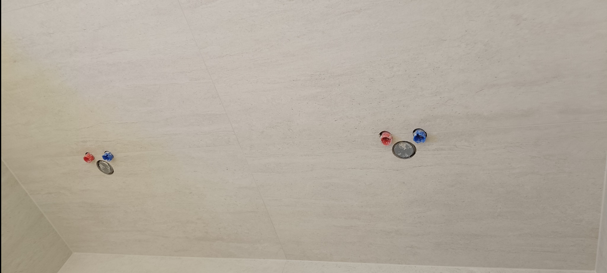 Jasne płytki imitujące kamień na ścianie z przygotowanymi otworami na baterię i przyłącza wody (czerwone i niebieskie). Widok z dołu.