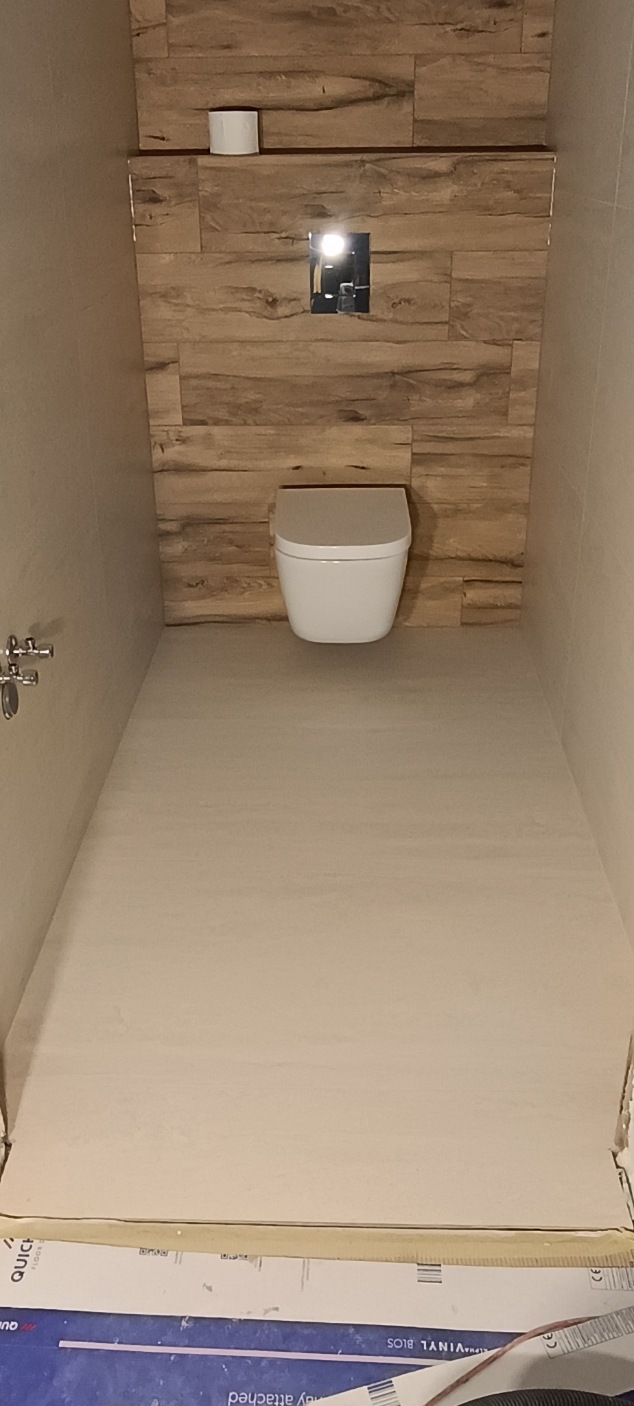 Nowoczesna toaleta podwieszana z drewnopodobną okładziną ścienną i minimalistycznym przyciskiem spłukującym. Jasna podłoga i neutralne barwy tworzą spójną całość.