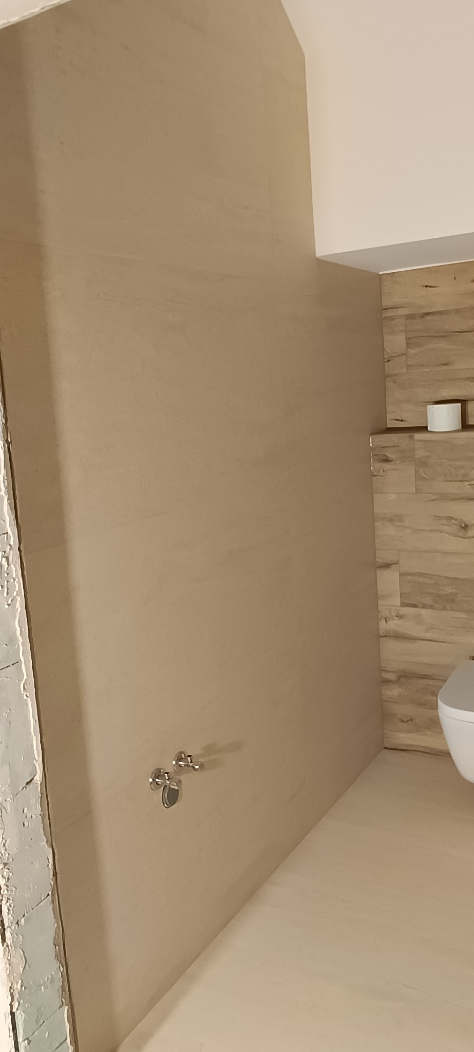 Wykończona łazienka z beżowymi płytkami imitującymi drewno na ścianie i podłodze. Widoczne chromowane uchwyty na ręczniki i fragment toalety. Ściana po lewej w trakcie wykończenia.