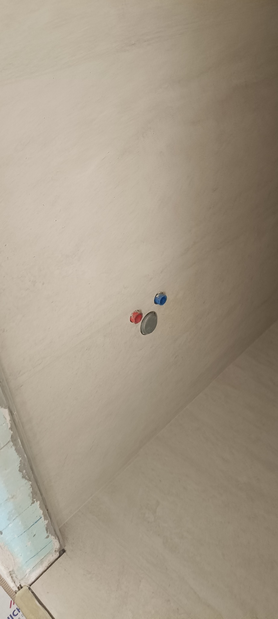 Ściana po nałożeniu gładzi, widoczne wyprowadzenia instalacji wodnej z zaślepkami w kolorze niebieskim i czerwonym oraz otwór kanalizacyjny.