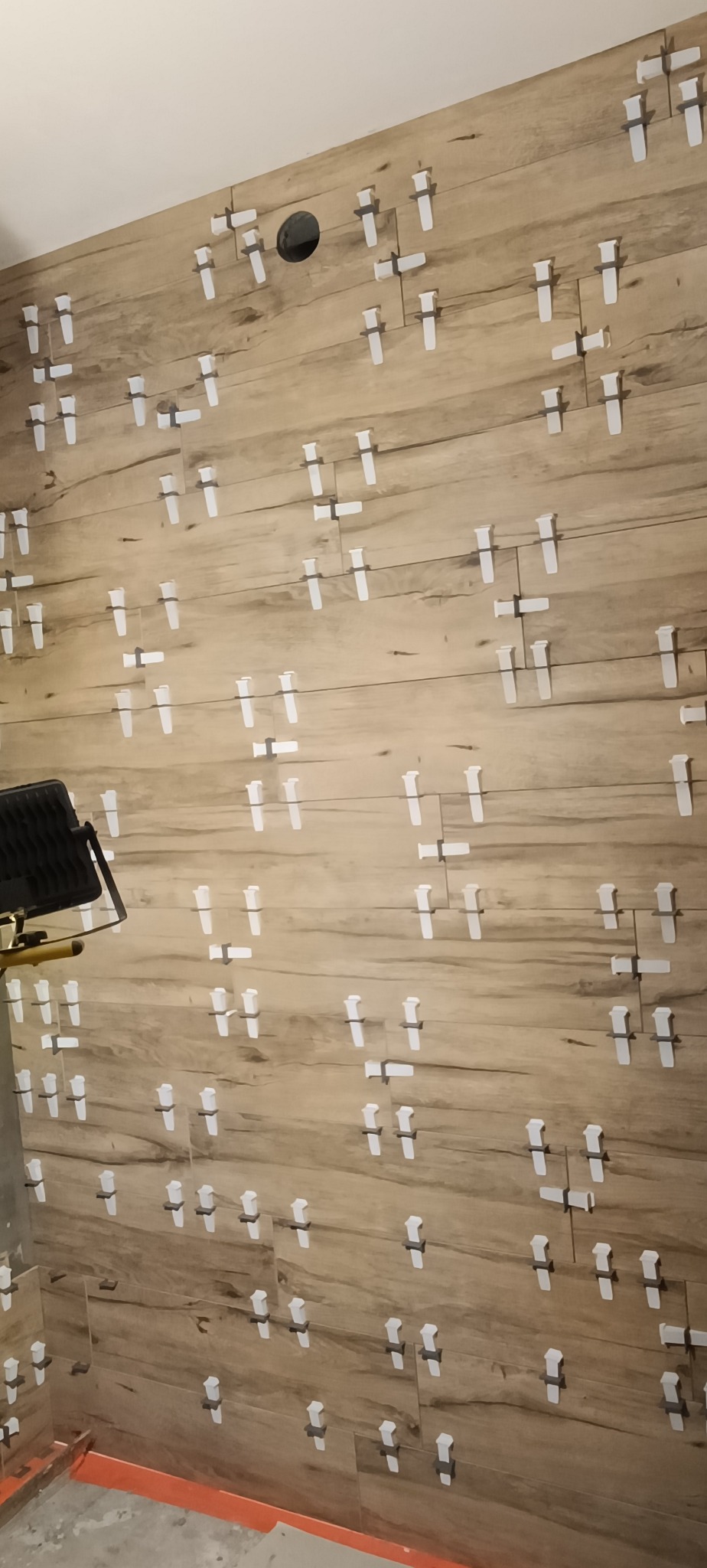 Ściana wyłożona płytkami imitującymi drewno, z użyciem białych krzyżyków dystansowych. Widoczny fragment lampy halogenowej doświetlającej prace.