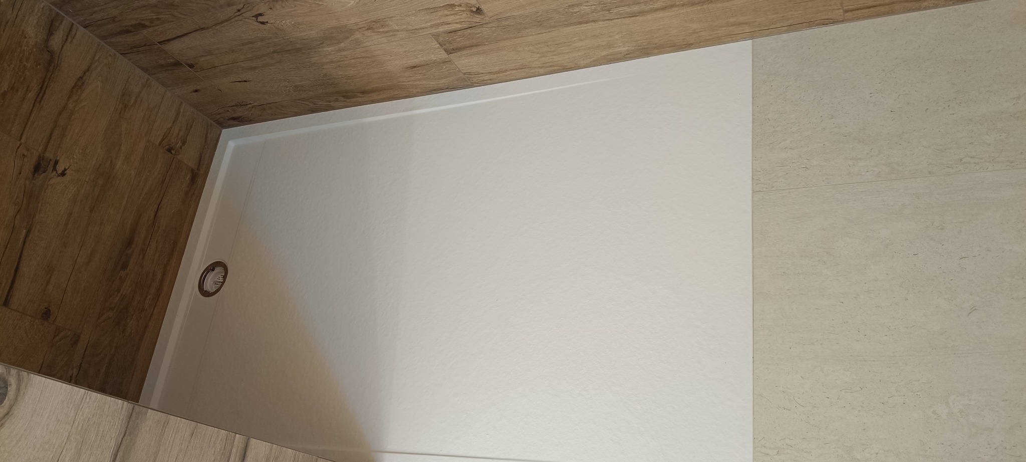 Biały brodzik z odpływem w narożnej kabinie prysznicowej, wykończonej płytkami imitującymi drewno i kamień. Minimalistyczny design.