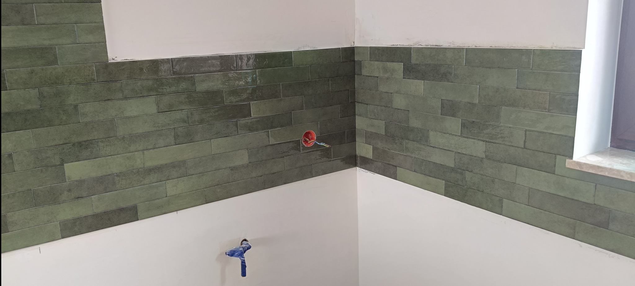 Ściana wyłożona podłużnymi, zielonymi płytkami w łazience, z widocznymi puszkami elektrycznymi i fragmentem okna. Płytki ułożone do pewnej wysokości, powyżej biała ściana.