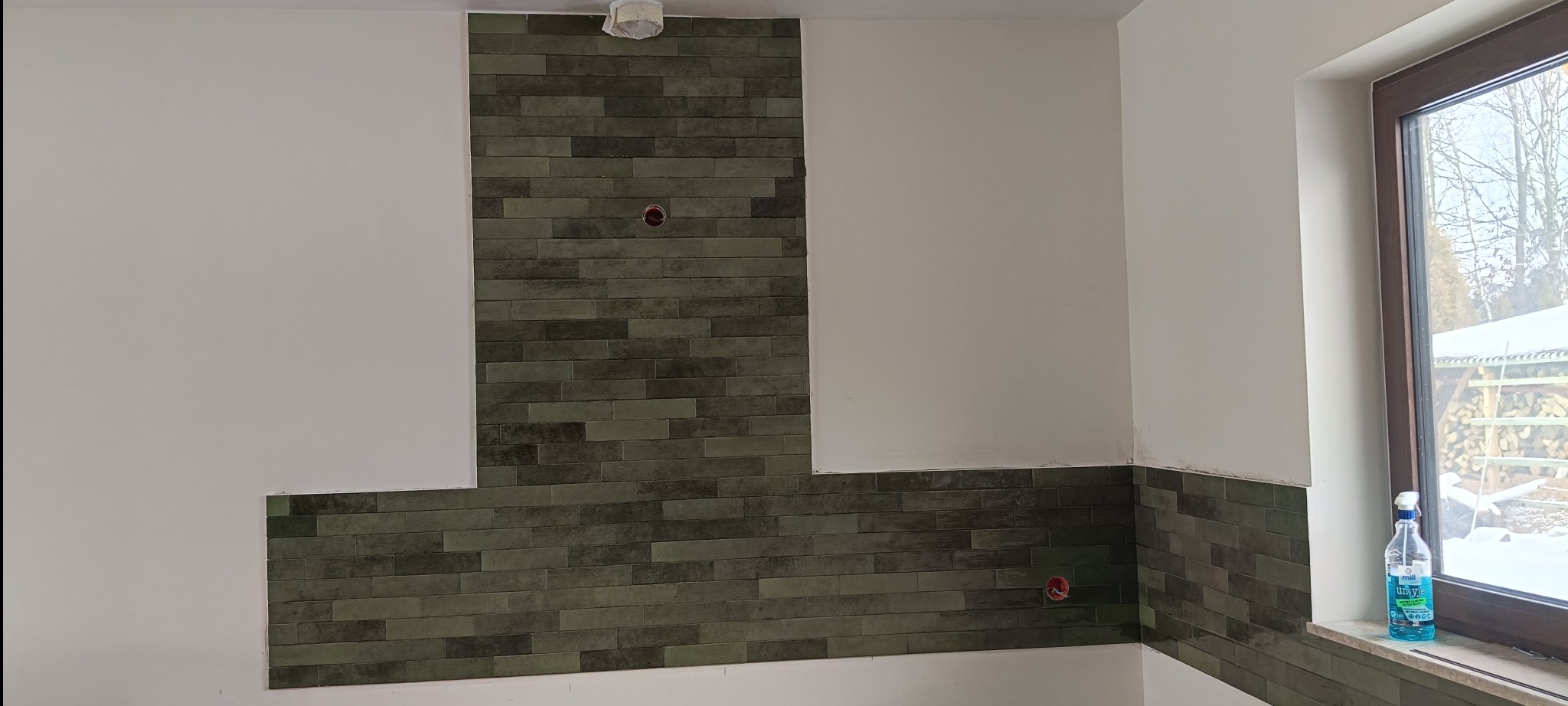 Fragment ściany z ułożoną, zielono-szarą, prostokątną glazurą, częściowo wykończoną, z widocznymi puszkami elektrycznymi i oknem z widokiem na zimowy krajobraz.