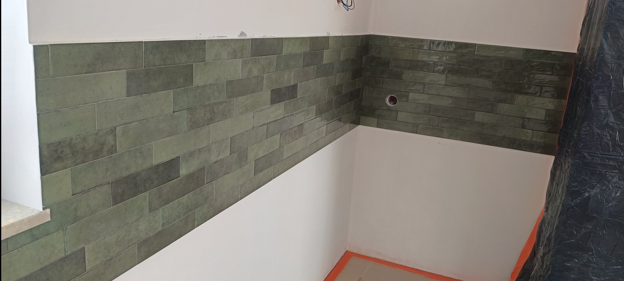 Ściana wyłożona podłużnymi, oliwkowymi płytkami ceramicznymi w połowie wysokości. Widoczne przygotowania do dalszych prac wykończeniowych, zabezpieczone folią.