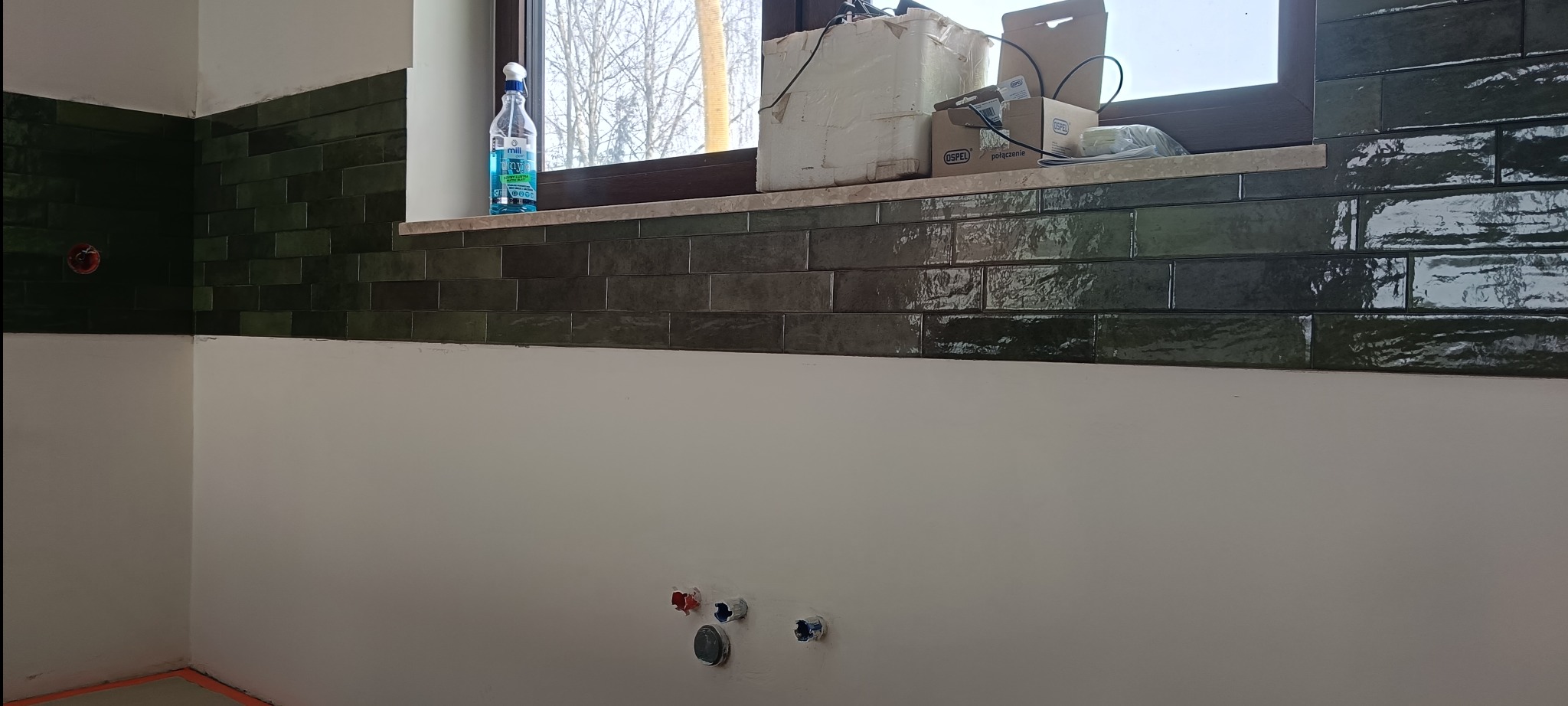 Położone, błyszczące, ciemnozielone płytki w kuchni, widoczne instalacje wodne w ścianie. Na parapecie okiennym kartony i butelka z płynem.
