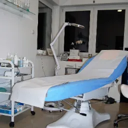 Kozetka kosmetyczna z regulowanym oparciem, nakryta białym prześcieradłem i niebieskim podkładem, obok wózek z kosmetykami i lampa powiększająca, w tle okno i półki z produktami.