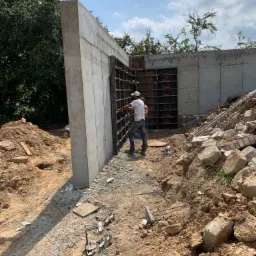 Budowa betonowej ściany fundamentowej z widocznym szalunkiem i dwoma pracownikami w tle, teren budowy z gruzem i ziemią.