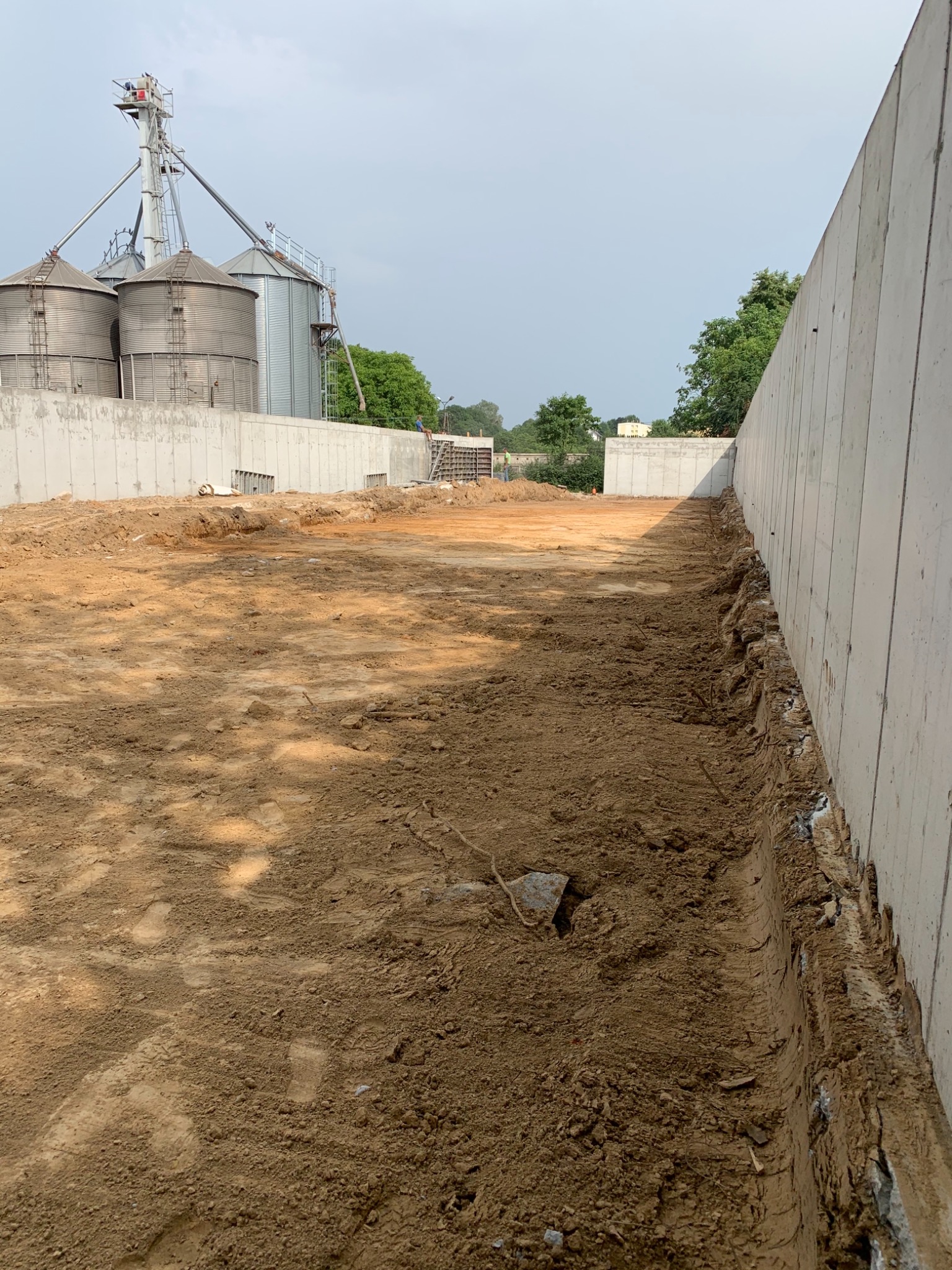 Wykop pod fundament z widocznymi ścianami oporowymi i silosami zbożowymi w tle.