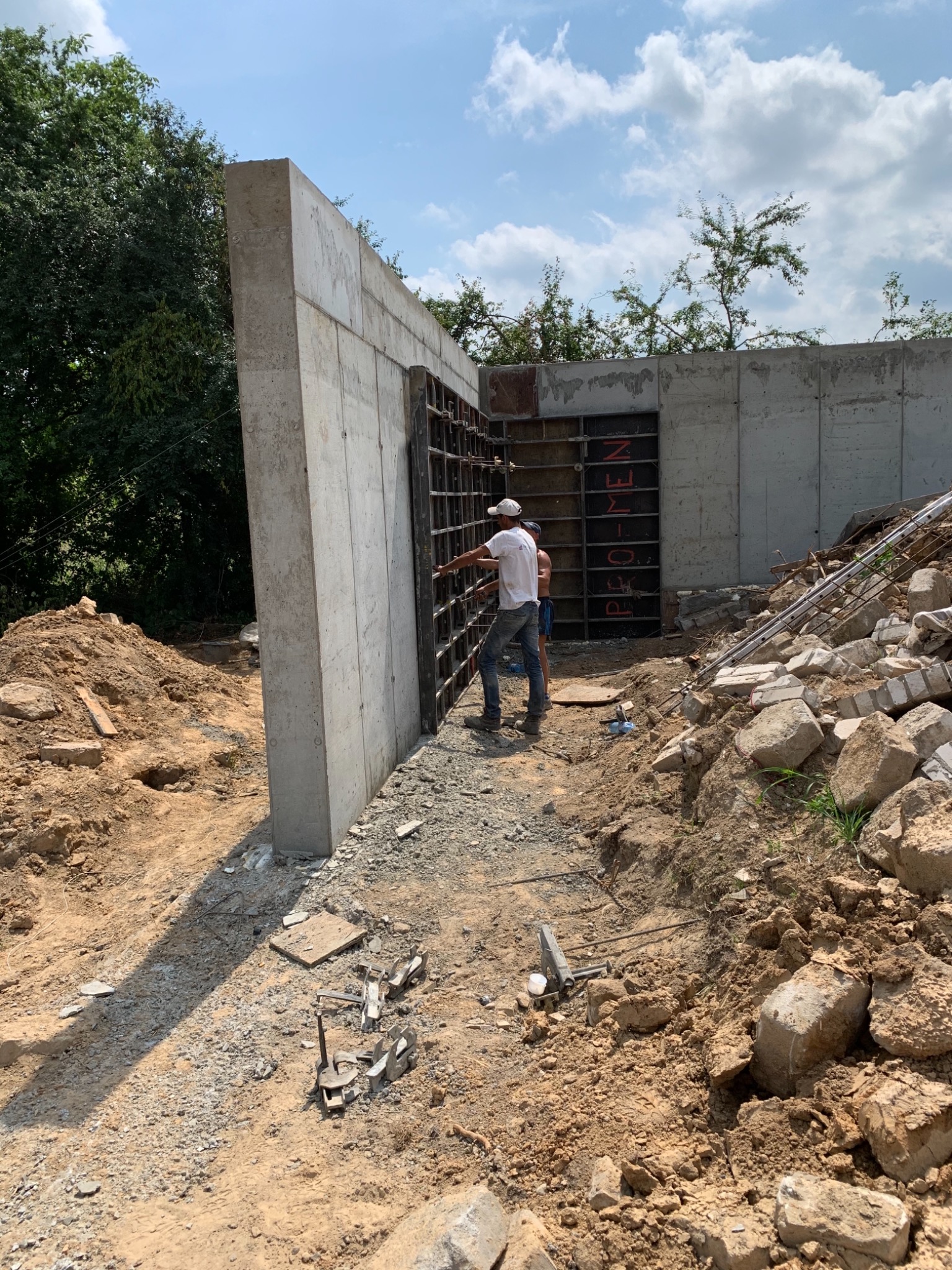 Budowa betonowej ściany fundamentowej z widocznym szalunkiem i dwoma pracownikami w tle, teren budowy z gruzem i ziemią.