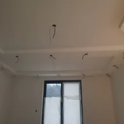 Biały sufit z elementami dekoracyjnymi i wystającymi kablami elektrycznymi, okno zasłonięte folią ochronną w tle.