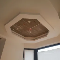 Wnętrze w trakcie remontu: sufit z widoczną betonową konstrukcją w kształcie ośmiokąta, obramowaną szarą listwą, okno zasłonięte folią malarską.