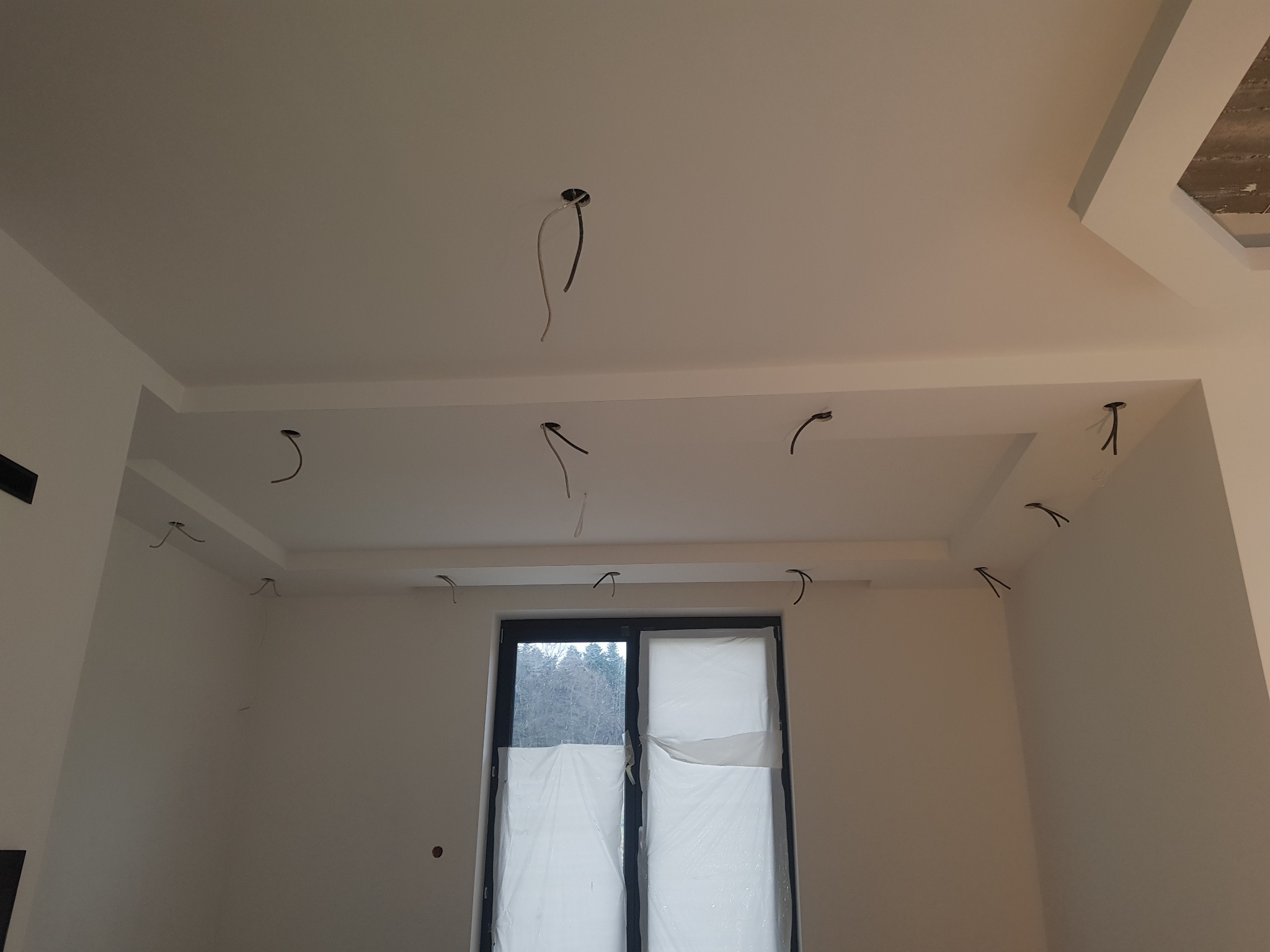 Biały sufit z elementami dekoracyjnymi i wystającymi kablami elektrycznymi, okno zasłonięte folią ochronną w tle.