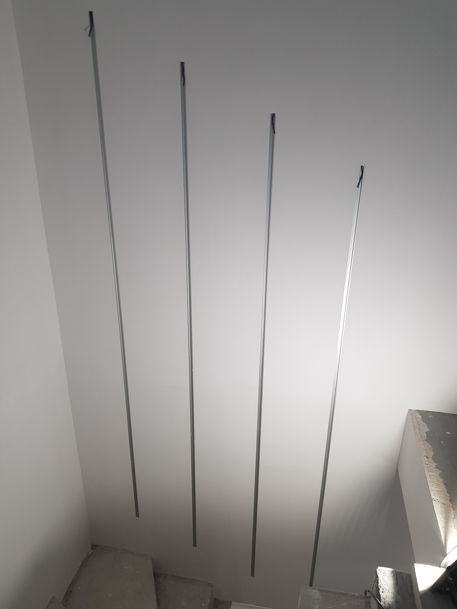 Metalowe profile zawieszone pionowo od sufitu, przygotowane do montażu konstrukcji podwieszanej, widoczne fragmenty betonowych schodów i surowy beton na ścianie.