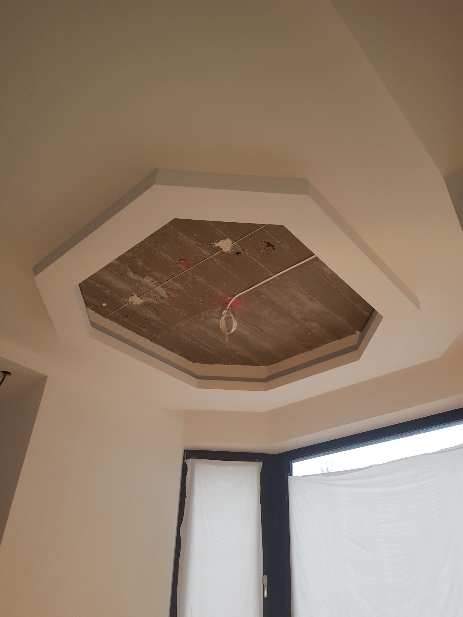 Wnętrze w trakcie remontu: sufit z widoczną betonową konstrukcją w kształcie ośmiokąta, obramowaną szarą listwą, okno zasłonięte folią malarską.