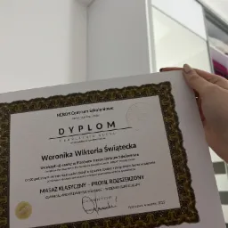 Dyplom ukończenia kursu masażu klasycznego z Heros Centrum Szkoleniowego, trzymany w dłoni na tle białej szafy przesuwnej.