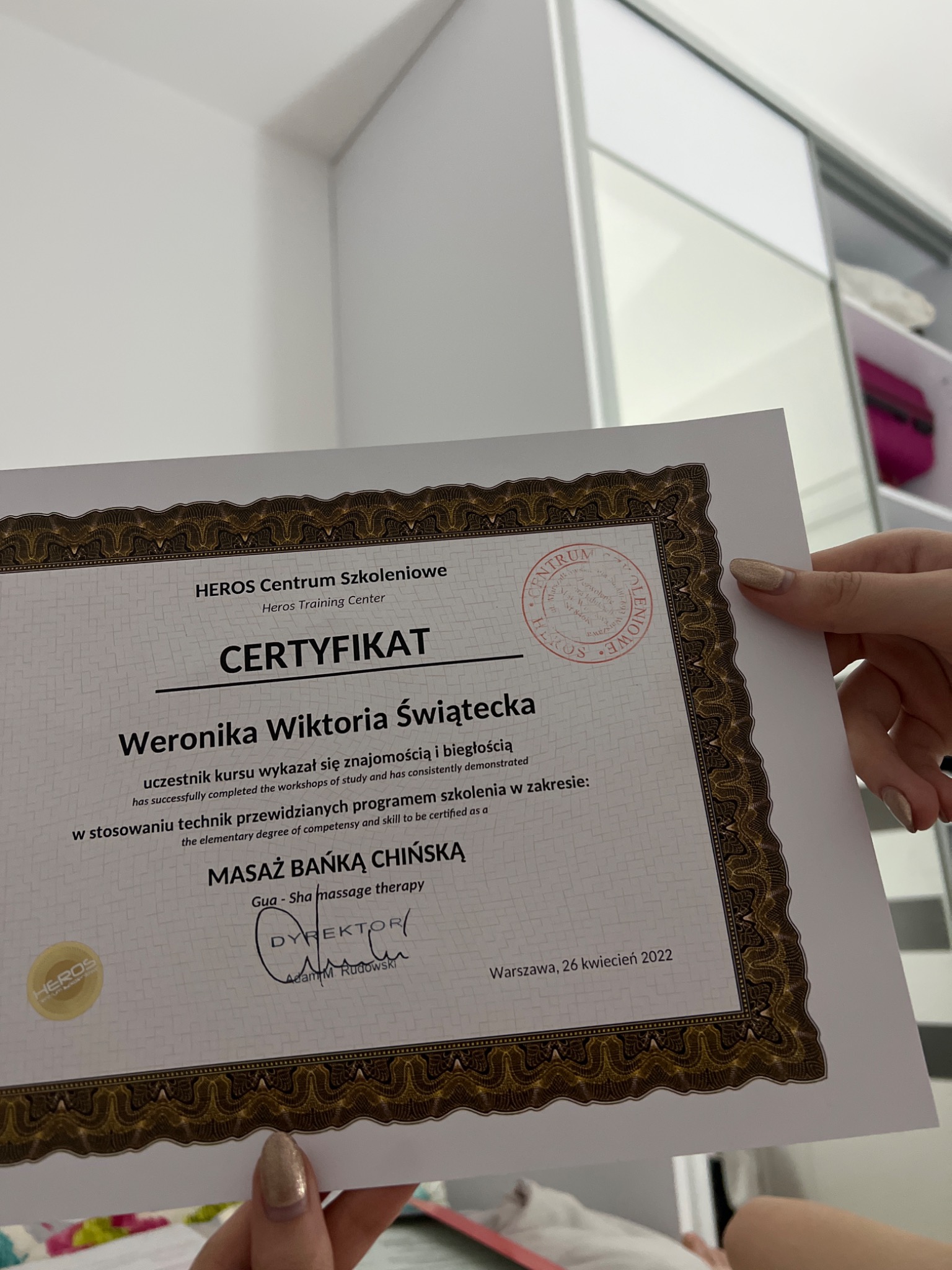 Certyfikat ukończenia kursu Masażu Bańką Chińską, wydany przez Heros Centrum Szkoleniowe dla Weroniki Wiktorii Świąteckiej, prezentowany na tle białej szafy.