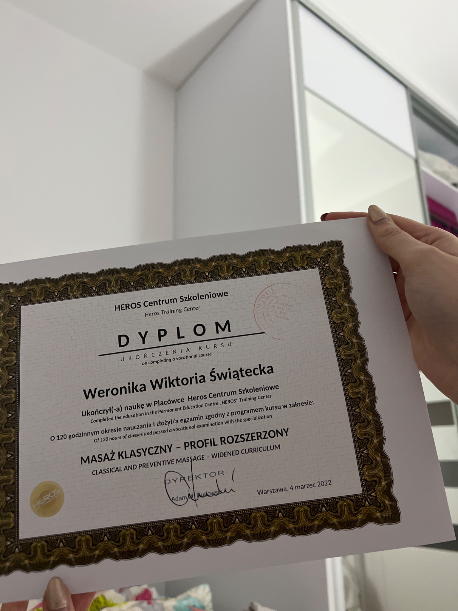 Dyplom ukończenia kursu masażu klasycznego z Heros Centrum Szkoleniowego, trzymany w dłoni na tle białej szafy przesuwnej.