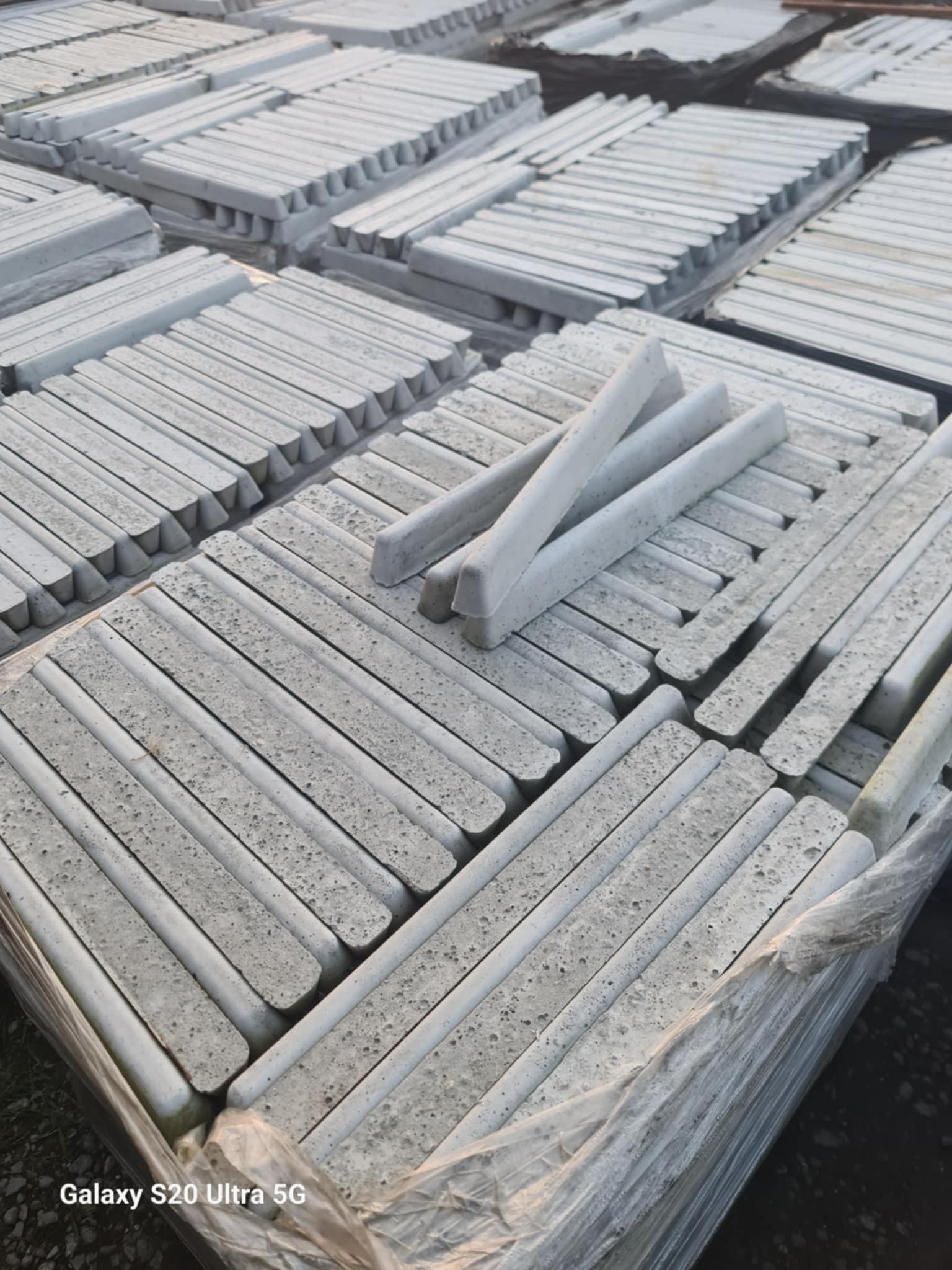 Szare obrzeża betonowe ułożone w stosy, widoczne pęknięcia i porowata struktura betonu. Trzy obrzeża ułożone na wierzchu stosu. Całość zapakowana w folię.