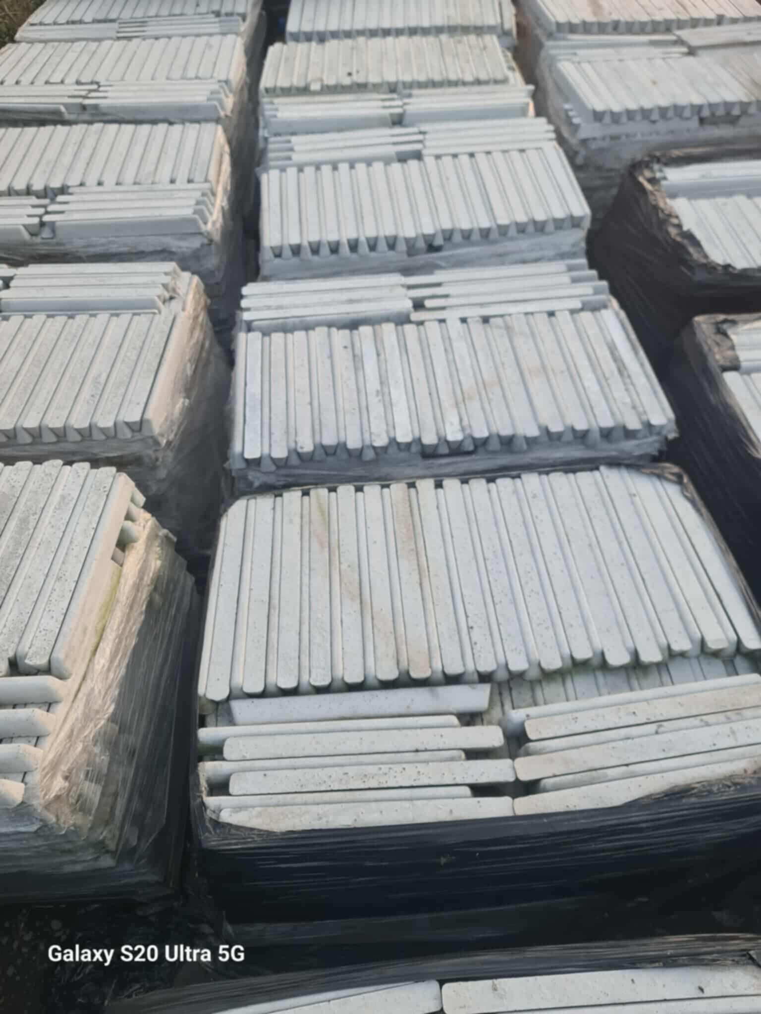 Stosy szarych, podłużnych prefabrykatów betonowych ułożonych w paczki, zabezpieczonych folią i czarną taśmą. Widoczne detale faktury betonu i krawędzi elementów.