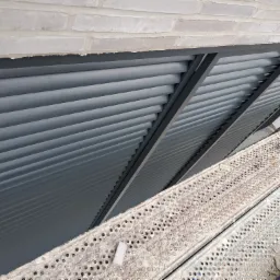 Szare, poziome lamele zewnętrzne zamontowane pod kątem, widoczne z rusztowania, z fragmentem ceglanego muru powyżej.