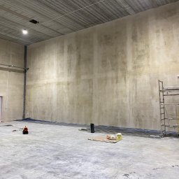 M-Bud - Wnętrze hali w trakcie przygotowania ścian do malowania: zabezpieczone podłoże, ustawiony ruszt, widoczny wałek malarski na rusztowaniu, wentylator osuszający w tle.