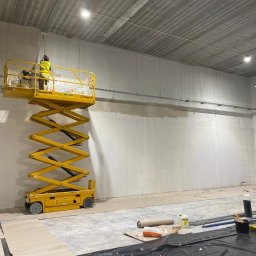 M-Bud - Pracownik na podnośniku nożycowym maluje ścianę wewnątrz hali, widoczna pierwsza warstwa świeżej farby, przygotowane narzędzia malarskie i folia ochronna na podłodze.