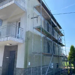 Dwupiętrowy budynek w trakcie czyszczenia elewacji, widoczne rusztowania i fragment ściany z zielonym nalotem przed usunięciem.