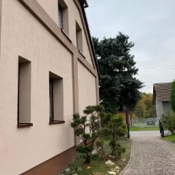 Odnowiona elewacja domu jednorodzinnego z brązowymi akcentami wokół okien i w dolnej części ściany, widoczna z boku, z zadbanym ogrodem i brukowaną ścieżką.