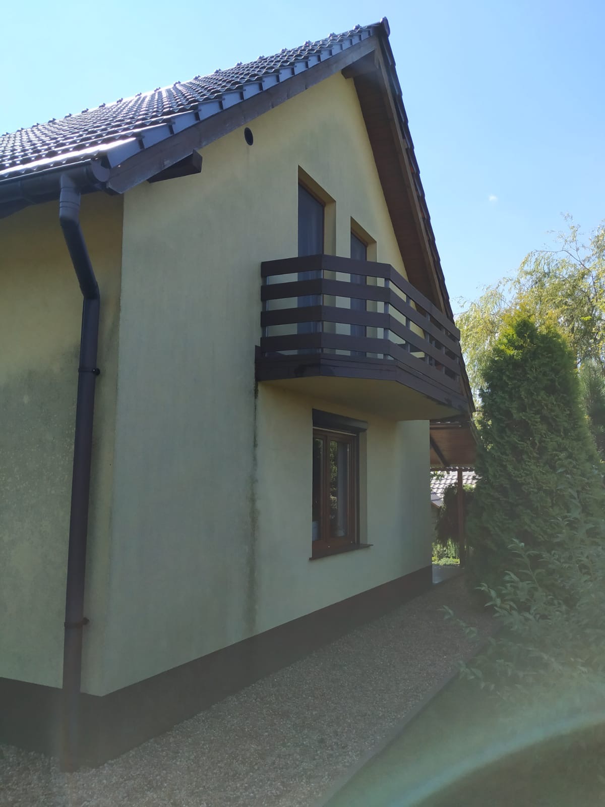 Żółta elewacja domu jednorodzinnego z brązowymi ramami okiennymi, balkonem i rynną, widoczne zabrudzenia na ścianie.