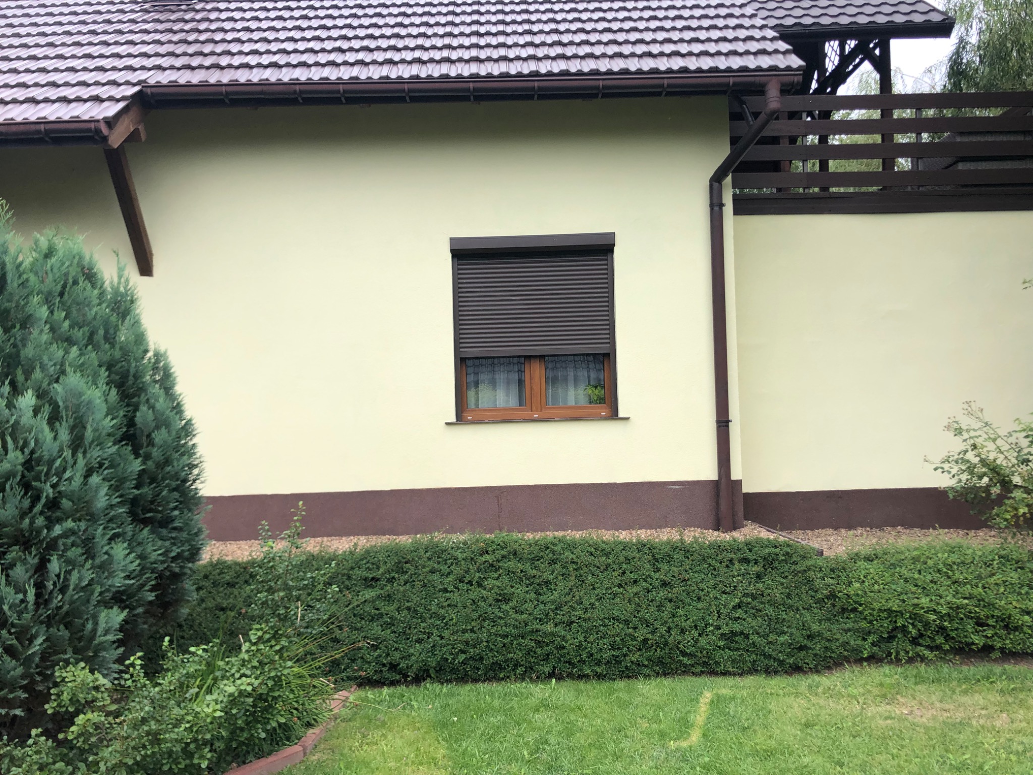 Żółta elewacja domu jednorodzinnego z brązowymi akcentami, brązowa roleta na oknie, zadbany trawnik i krzewy przed budynkiem.