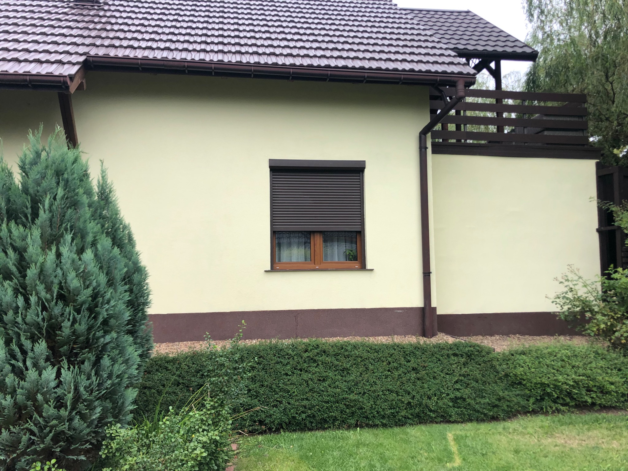 Elewacja domu jednorodzinnego z zamkniętą roletą zewnętrzną, widoczny fragment balkonu, brązowy cokół i zielone krzewy w ogrodzie.