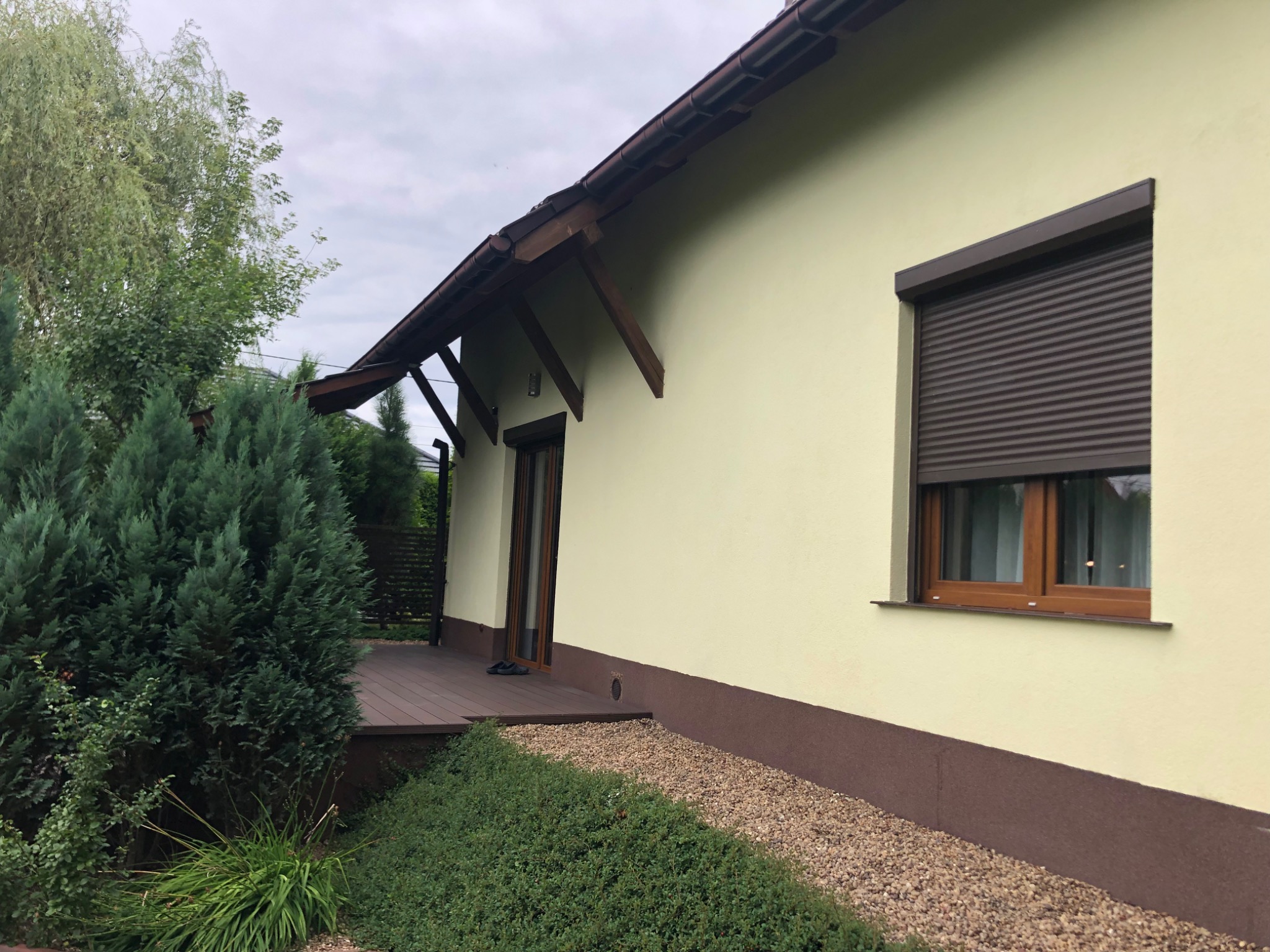 Jasnożółta elewacja domu jednorodzinnego z brązowymi elementami wykończeniowymi, widoczne rolety zewnętrzne, drewniany taras i ozdobne krzewy.