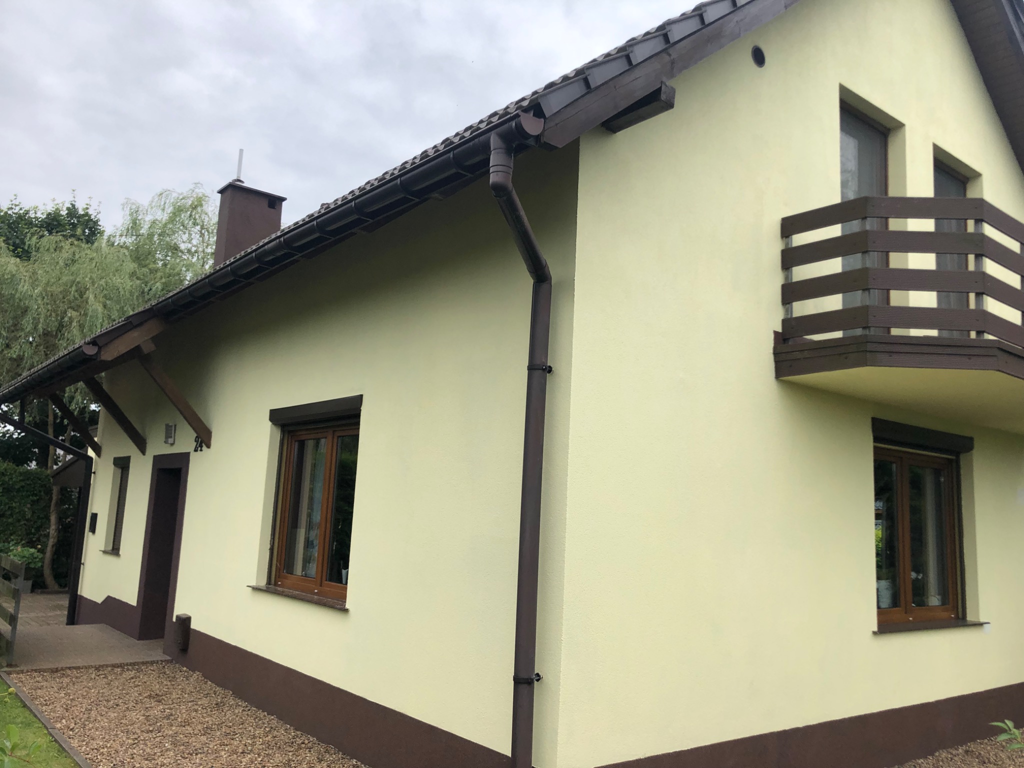 Jasnożółty dom jednorodzinny z brązowymi elementami wykończeniowymi: podmurówka, okna, rynny, dach i balkon z drewnianą balustradą, widok z zewnątrz w pochmurny dzień.