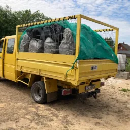 Żółta ciężarówka z otwartą skrzynią ładunkową wypełnioną workami z odpadami, zabezpieczona zieloną siatką, zaparkowana na piaszczystym terenie.