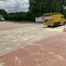 Żółta ciężarówka z piaskiem na pace stoi na parkingu z kostki, obok widoczny paczkomat i zieleń drzew.