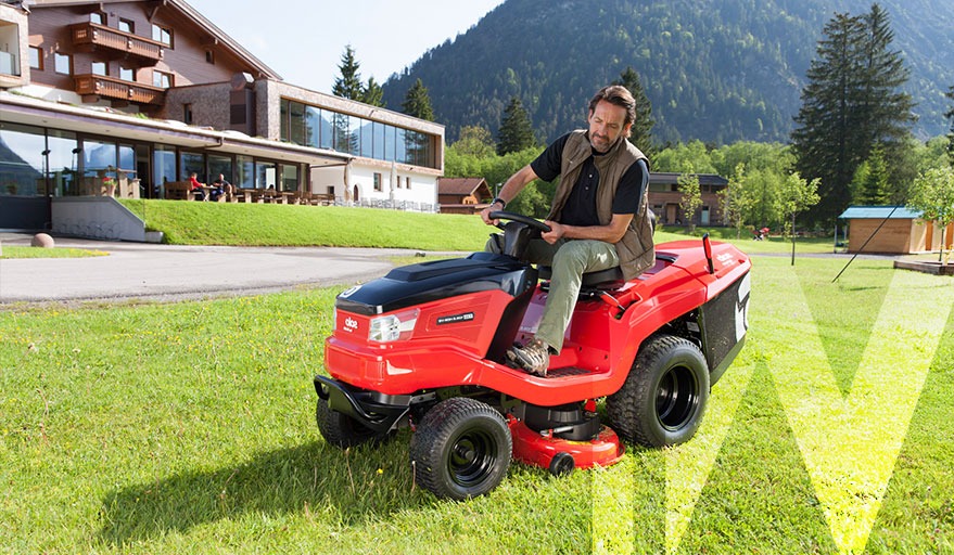 Mężczyzna prowadzi czerwoną kosiarkę traktorek na trawniku przed budynkiem z drewnianymi balkonami i widokiem na góry.