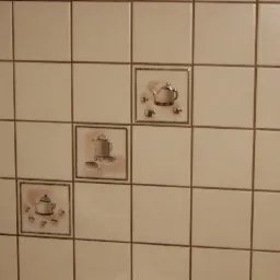 Ściana wyłożona kwadratowymi, beżowymi płytkami ceramicznymi z dekoracyjnymi wstawkami przedstawiającymi motywy czajników i filiżanek.