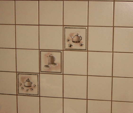 Ściana wyłożona kwadratowymi, beżowymi płytkami ceramicznymi z dekoracyjnymi wstawkami przedstawiającymi motywy czajników i filiżanek.